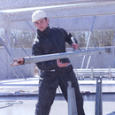 A construction worker wearing a black Lyngsoe LR1389 Rainset Jacket and Trousers lifts a heavy metal bar on a rainy construction site. The rain gear appears to be effectively repelling the water, keeping the worker dry and comfortable despite the wet conditions.