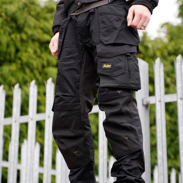 Lifestyle image showing a worker wearing black Snickers work trousers with stretch fabric, reinforced knee panels and utility pockets, photographed outdoors with a metal fence and greenery in the background, used as the main visual for a Best of Snickers Workwear 2026 blog post.
