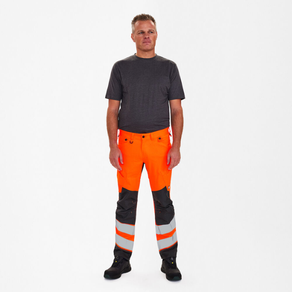 Safety pants worn by a model, front facing, in fluorescent orange with grey contrast and reflective bands.