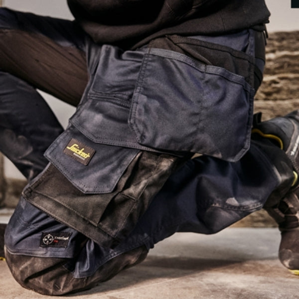 Close‑up view of Snickers 6241 work trousers showing the reinforced pockets, knee pad section and FlexiWork fabric while in a kneeling position on a work surface, highlighting the construction‑ready design and details for the Snickers 6241 collection.